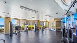 Un gimnasio con equipo de ejercicio diverso, ventanas amplias y vistas a edificios.