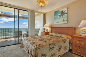 Una habitación de hotel con una cama, una mesita de noche con lámpara, un cuadro de una palmera y un balcón con vista al océano.