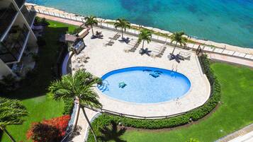 Luftaufnahme eines Resorts mit einem kreisförmigen Pool, umgeben von Liegestühlen und Palmen, mit Blick auf einen Strand.