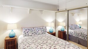 A bedroom with a large bed, two bedside tables with lamps, a mirror, and a wooden floor. - Wavecrest (Kaunakakai)