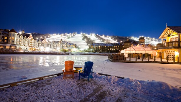 A snow-covered waterfront with two Adirondack chairs, a wooden dock, and a row of illuminated buildings in the background. - Snowbridge (The Blue Mountains)