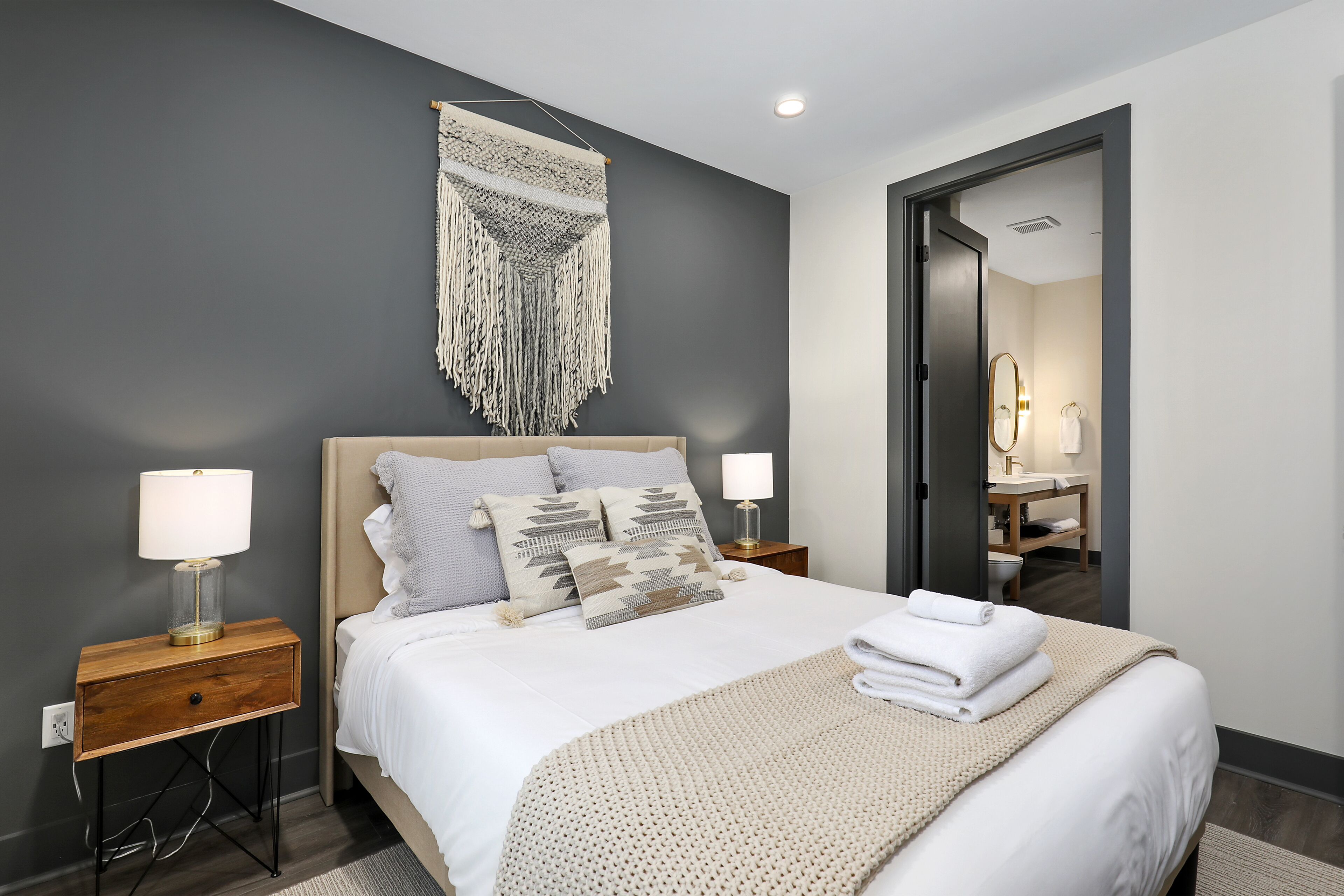 Une chambre d’hôtel moderne avec un grand lit, deux tables de chevet avec des lampes, une décoration murale en macramé et un miroir dans la salle de bain.