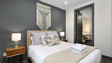 Une chambre d’hôtel moderne avec un grand lit, deux tables de chevet avec des lampes, une décoration murale en macramé et un miroir dans la salle de bain.