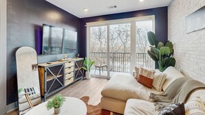A modern living room with a sofa, coffee table, and a large mirror. There is a sliding glass door leading to a balcony with a view of trees. - Muse Nashville (Nashville)
