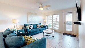 A living room with a blue sofa set, a round coffee table, a ceiling fan, and a door with a window. - Suntide II Condominiums (South Padre Island)