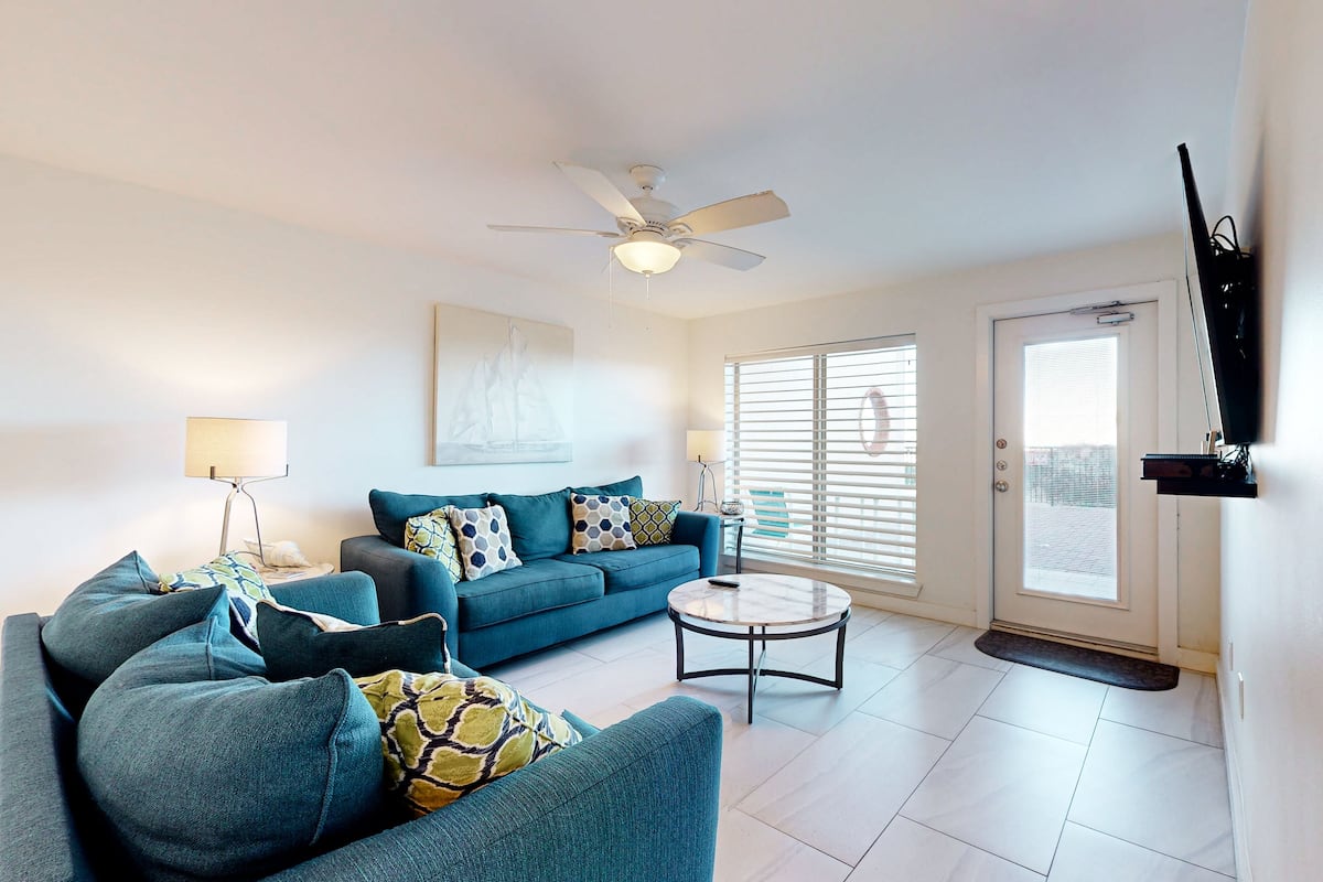 A living room with a blue sofa set, a round coffee table, a ceiling fan, and a door with a window.