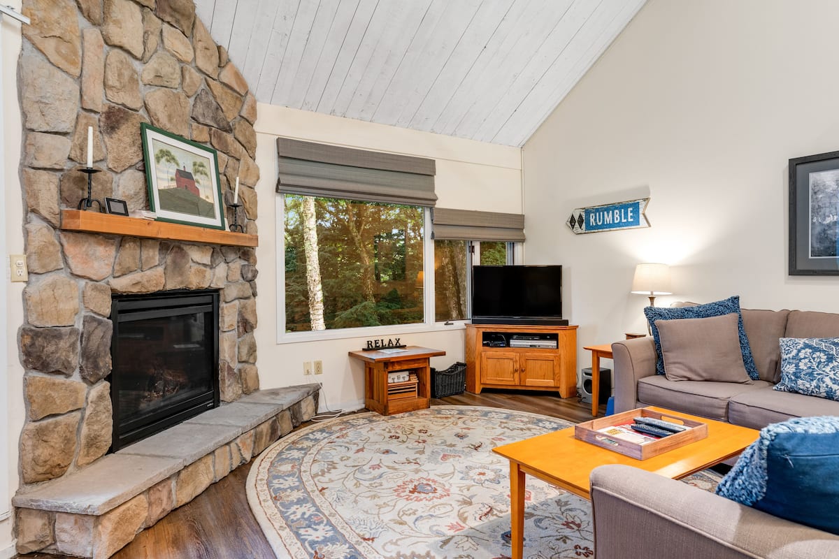 A cozy living room with a stone fireplace, a flat-screen TV, a comfortable sofa, and a large window offering a view of trees.