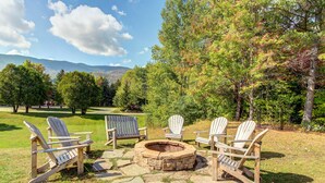 A stone fire pit surrounded by Adirondack chairs in a grassy outdoor area with trees and mountains in the background. - Bridges Resort (Warren)