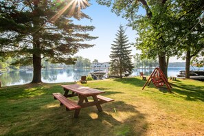 A lakeside picnic area with a wooden picnic table and benches, surrounded by trees and a swing set. - Thunderbird Resort Condo (Wisconsin Dells)