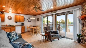 A cozy living room with a sofa, a chair, and a dining area with a table and chairs. There is a kitchen with wooden cabinets and a ceiling fan. A large window offers a view of a lake. - Thunderbird Resort Condo (Wisconsin Dells)