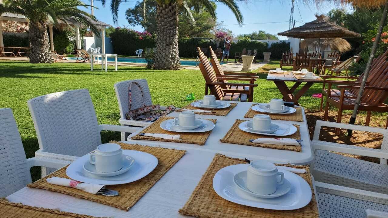 An outdoor dining area with white wicker chairs and tables set with white plates and cups, surrounded by lush greenery and a pool.