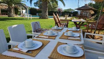 An outdoor dining area with white wicker chairs and tables set with white plates and cups, surrounded by lush greenery and a pool.