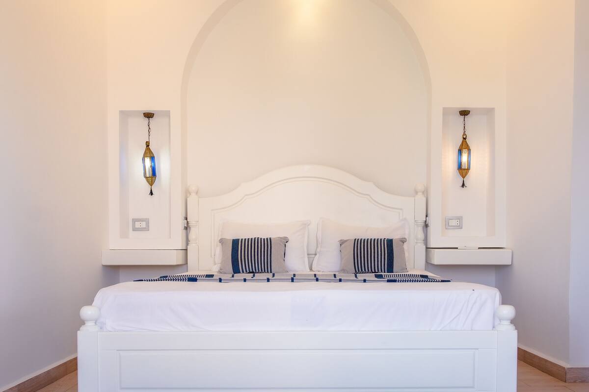 Een wit bed met een gebogen hoofdbord, twee nachtkastjes met lampen, en een witte muur met een gewelfde plafond.