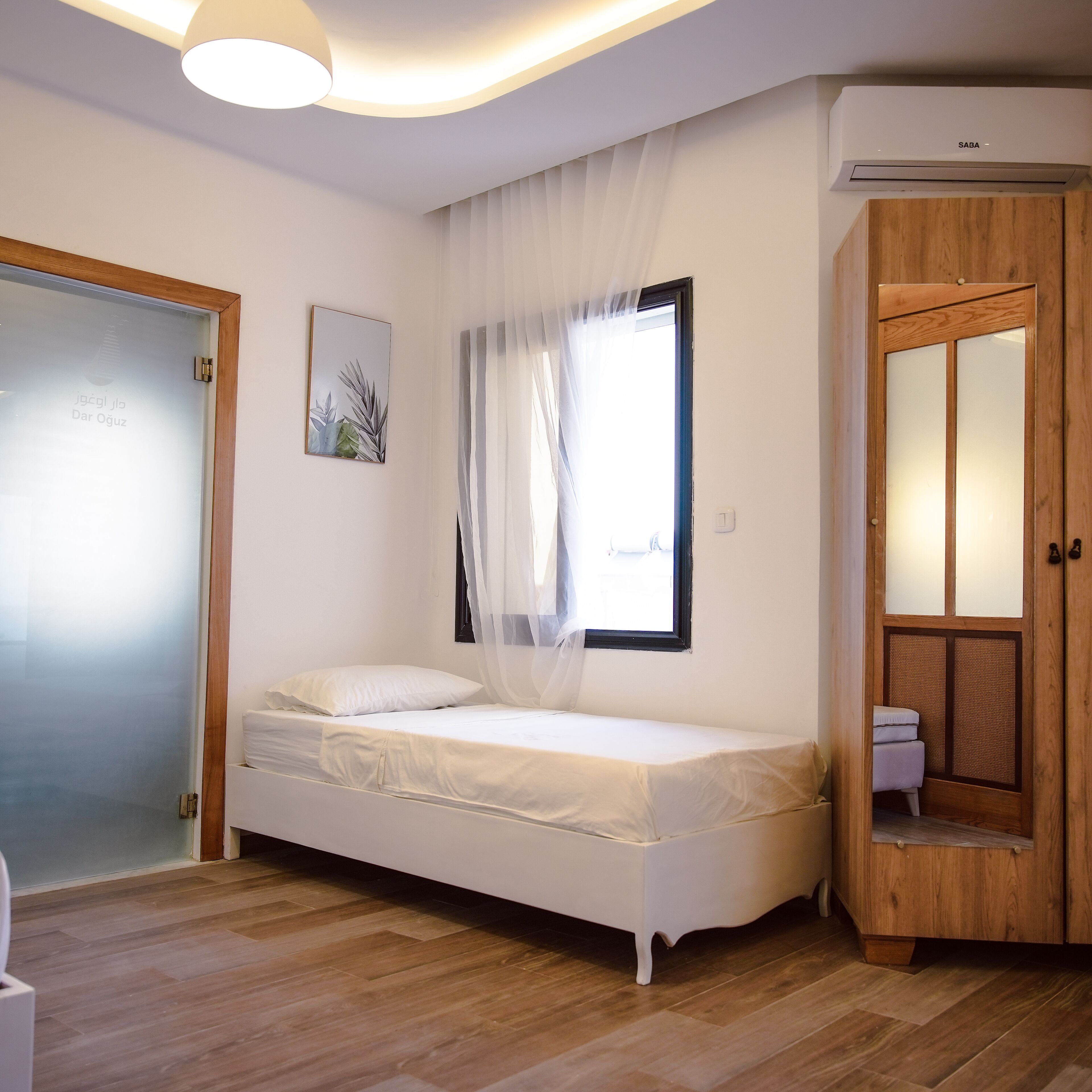 Un dormitorio con una cama individual, una ventana con cortinas traslúcidas, un armario de madera y un equipo de aire acondicionado instalado en la pared.
