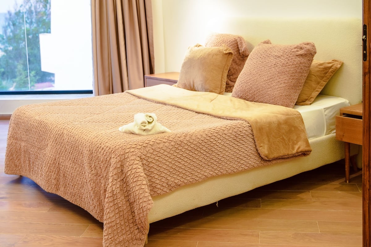 Een net opgemaakt bed met een gebreid plaid, een nachtkastje en een raam met gordijnen.