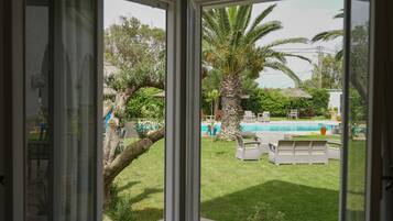A view from a room with a large window overlooking a backyard with a swimming pool, palm trees, and outdoor furniture.