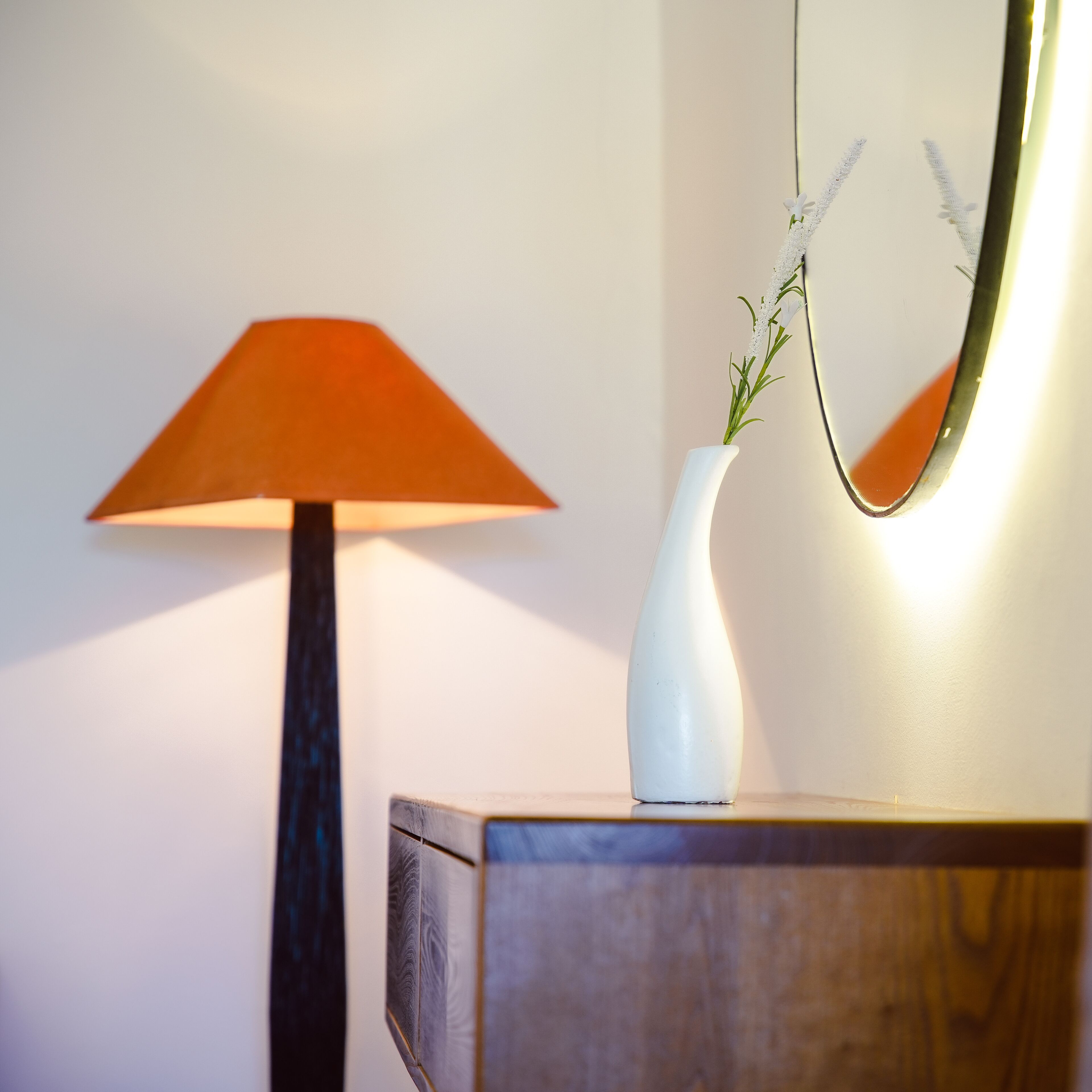Eine Lampe mit konischem Lampenschirm, eine weiße Vase mit Grünpflanzen und ein Spiegel auf einer Holzkommode vor einer schlichten Wand.