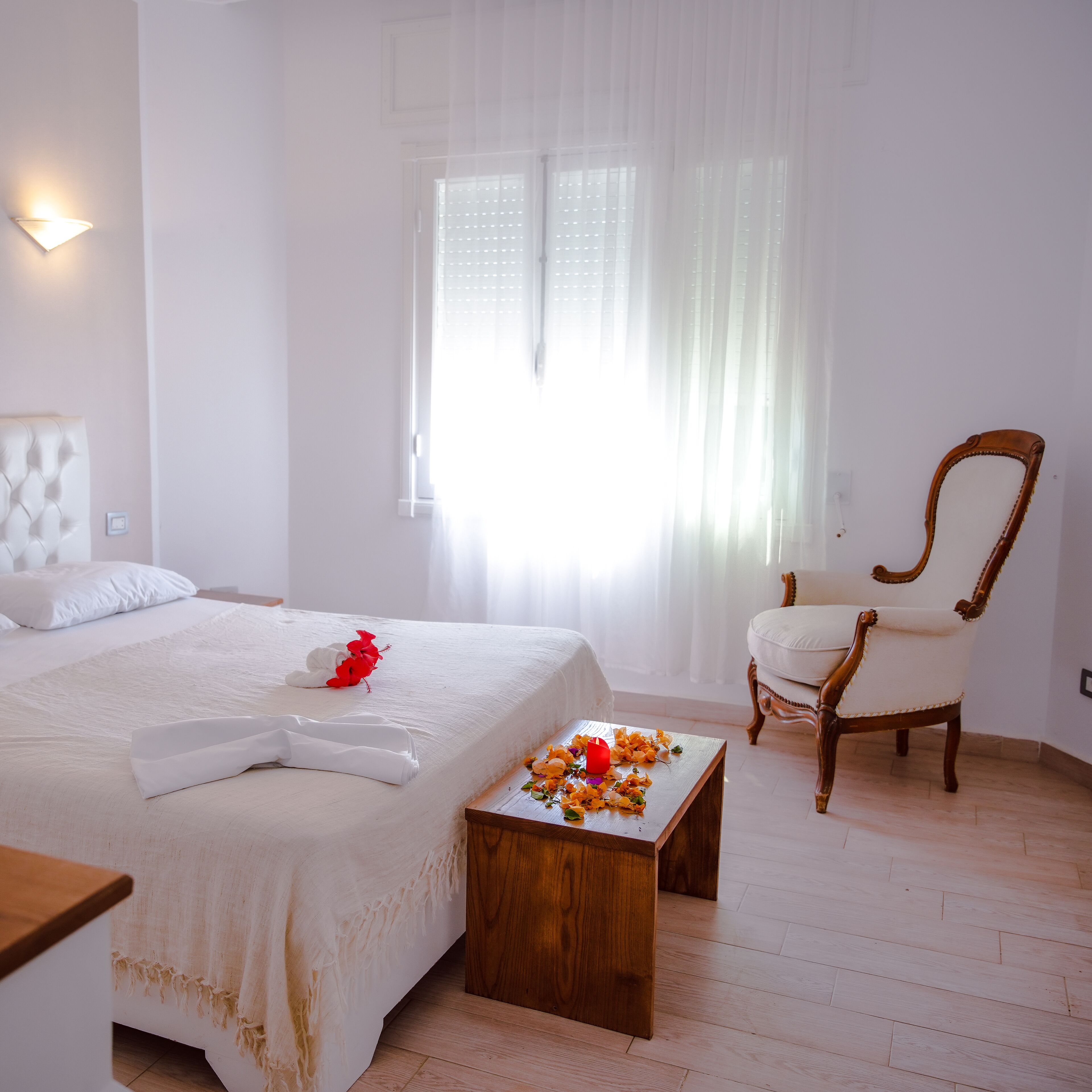 Una habitación de hotel con una cama, una mesita de madera con flores, una silla y una ventana con cortinas traslúcidas.