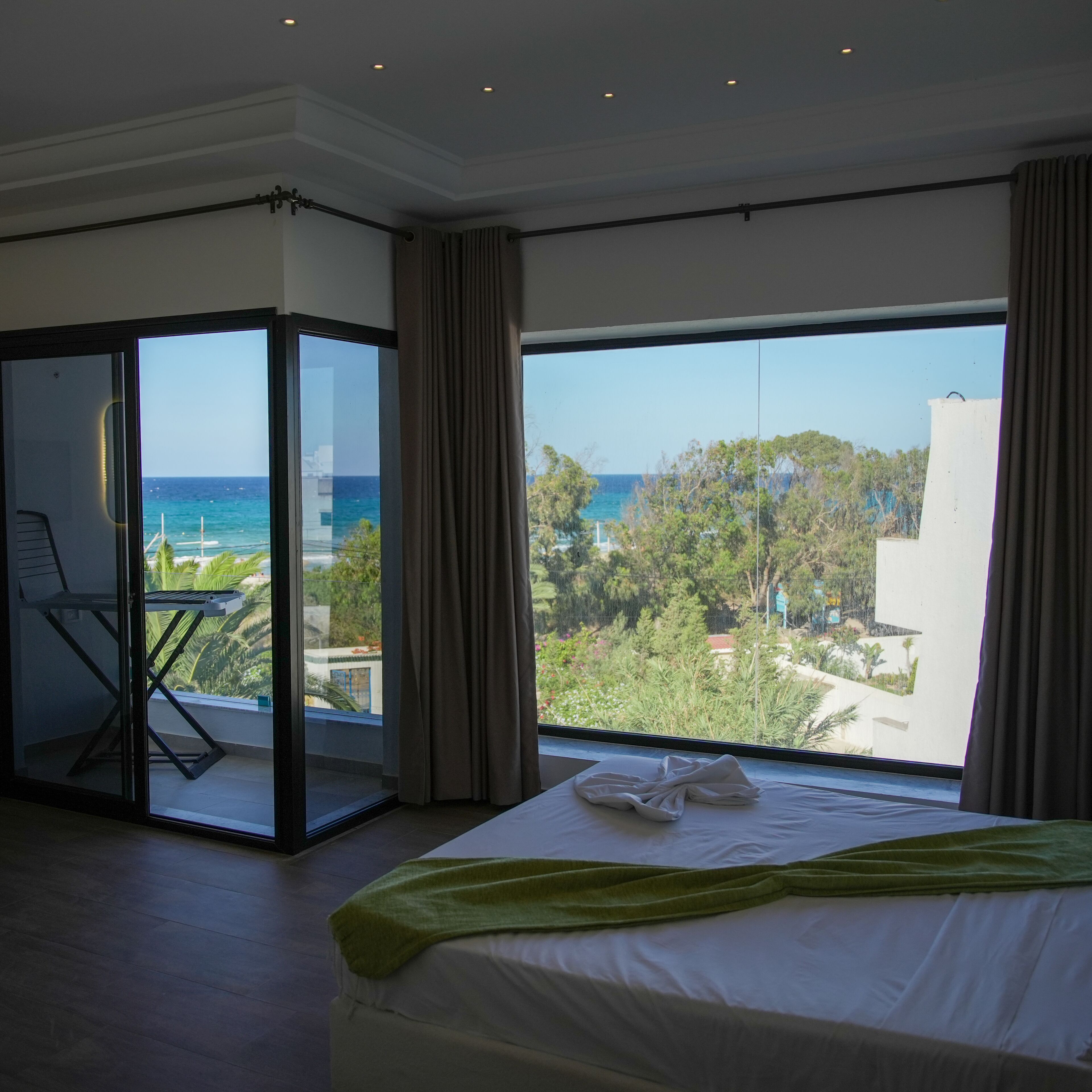 A hotel room with a large bed, a balcony with a view of the sea, and a sliding glass door leading to an outdoor area.