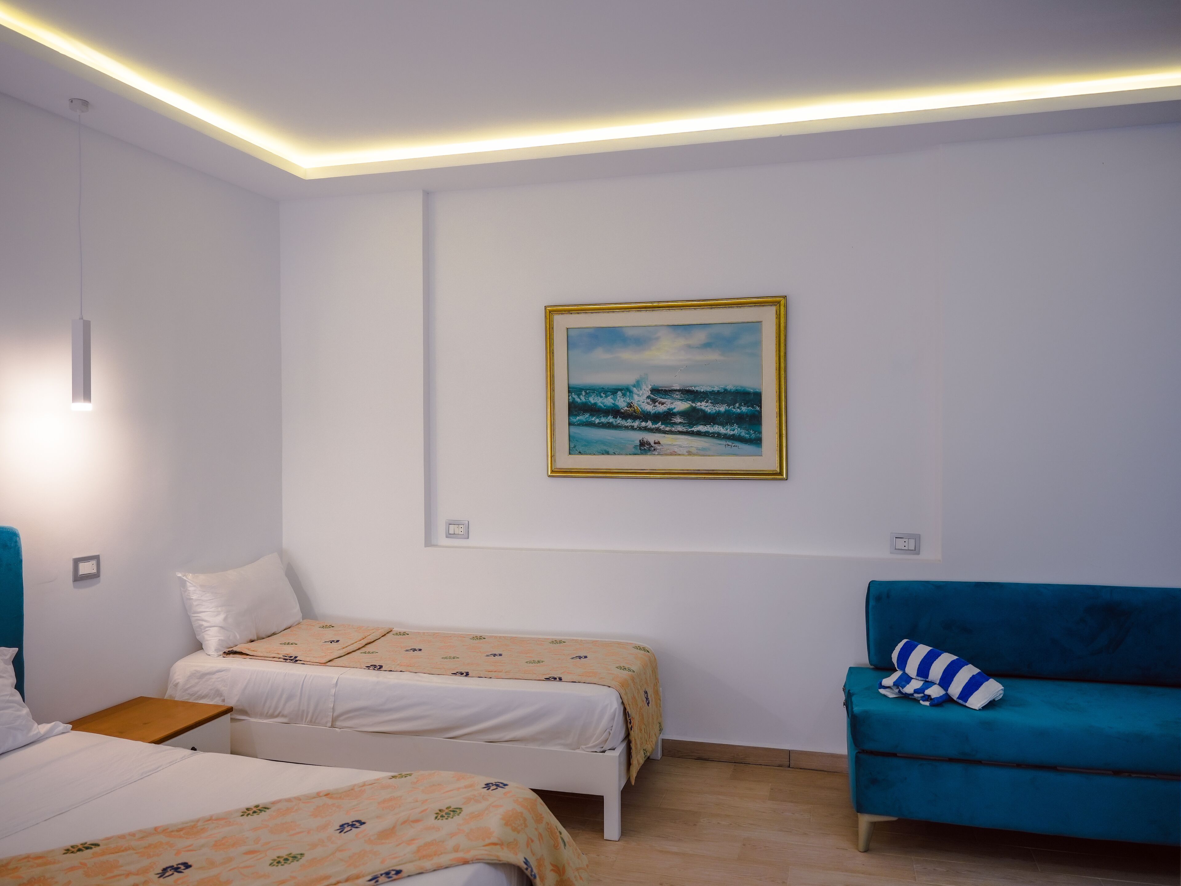 Una habitación de hotel con dos camas, un sofá azul, un cuadro de una escena marina y una lámpara en el techo.