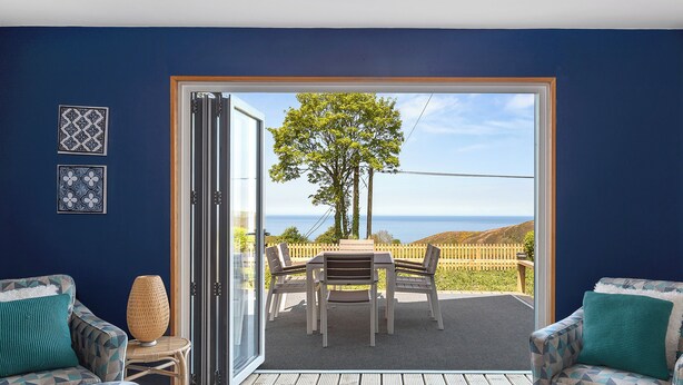 Una habitación interior con una puerta corrediza de vidrio que da a un patio exterior con mesa y sillas, vista al mar y decoración de pared con diseños.