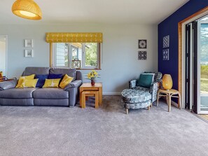 Un salón con un sofá gris, un sillón estampado, una mesita de madera y una ventana con persianas amarillas.