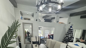 A modern dining area with a long table, white upholstered chairs, a chandelier, and a Christmas tree.