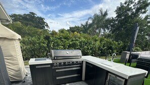 An outdoor kitchen with a grill, sink, and storage drawers, surrounded by a paved patio and greenery.