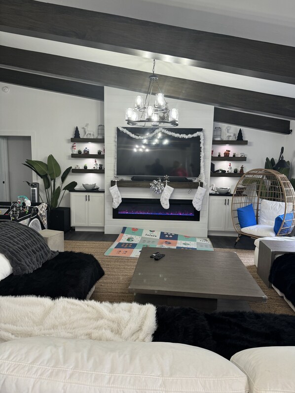 A modern living room with a fireplace, a chandelier, and a television. There are shelves with decorative items and a wicker chair. - JUST IN TIME FOR SEASON! (Bonita Springs)