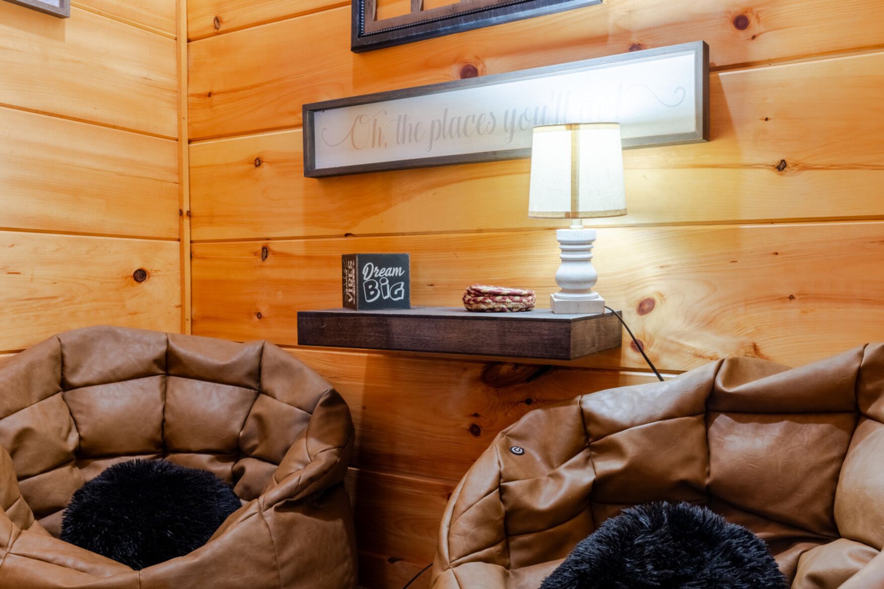 A cozy room with wooden walls, two leather armchairs with fluffy pillows, a small shelf with a sign, and a lamp.