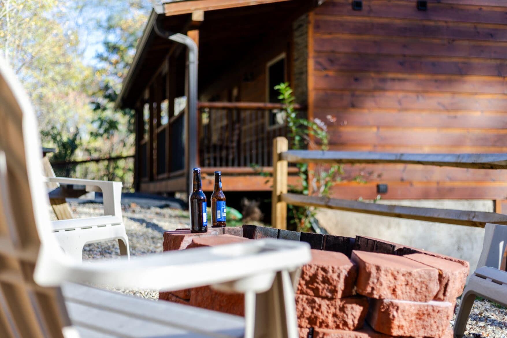 A wooden cabin with a porch, two bottles on a brick table, and white chairs.