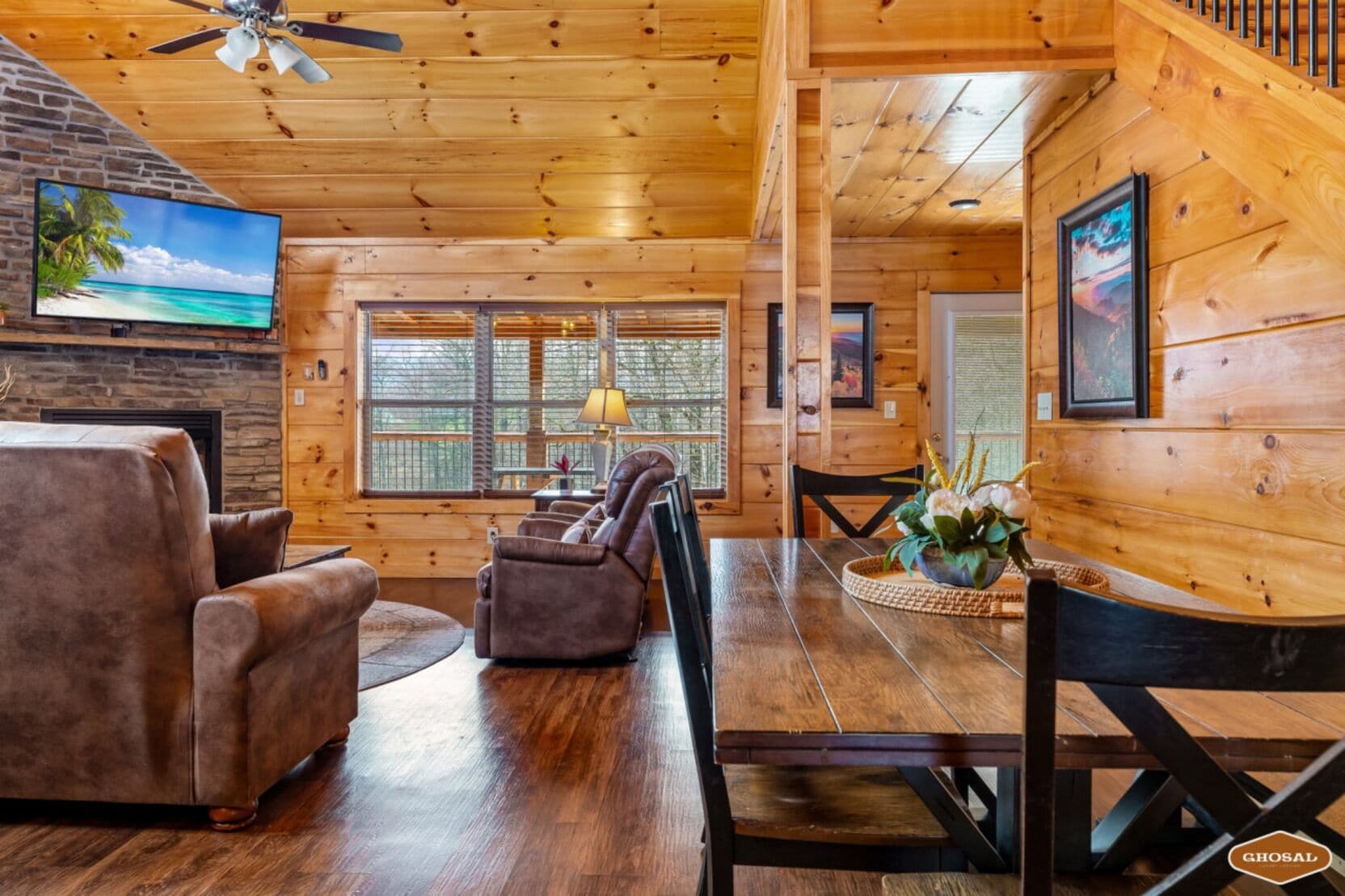 A wooden cabin interior with a fireplace, leather recliners, a dining table, and a TV mounted on the wall.