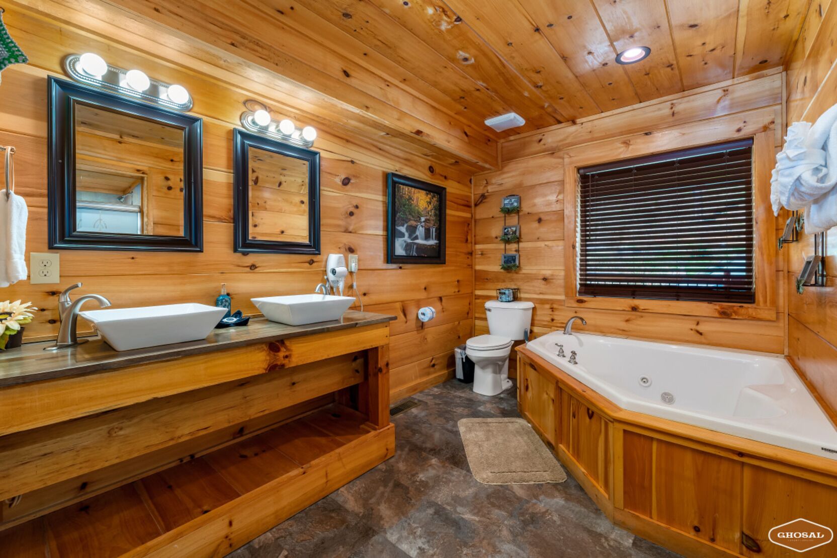 Une salle de bain rustique, avec des murs en bois, comprenant deux lavabos, des toilettes, une baignoire et une fenêtre équipée de stores.