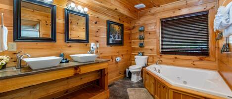 Une salle de bain rustique, avec des murs en bois, comprenant deux lavabos, des toilettes, une baignoire et une fenêtre équipée de stores.