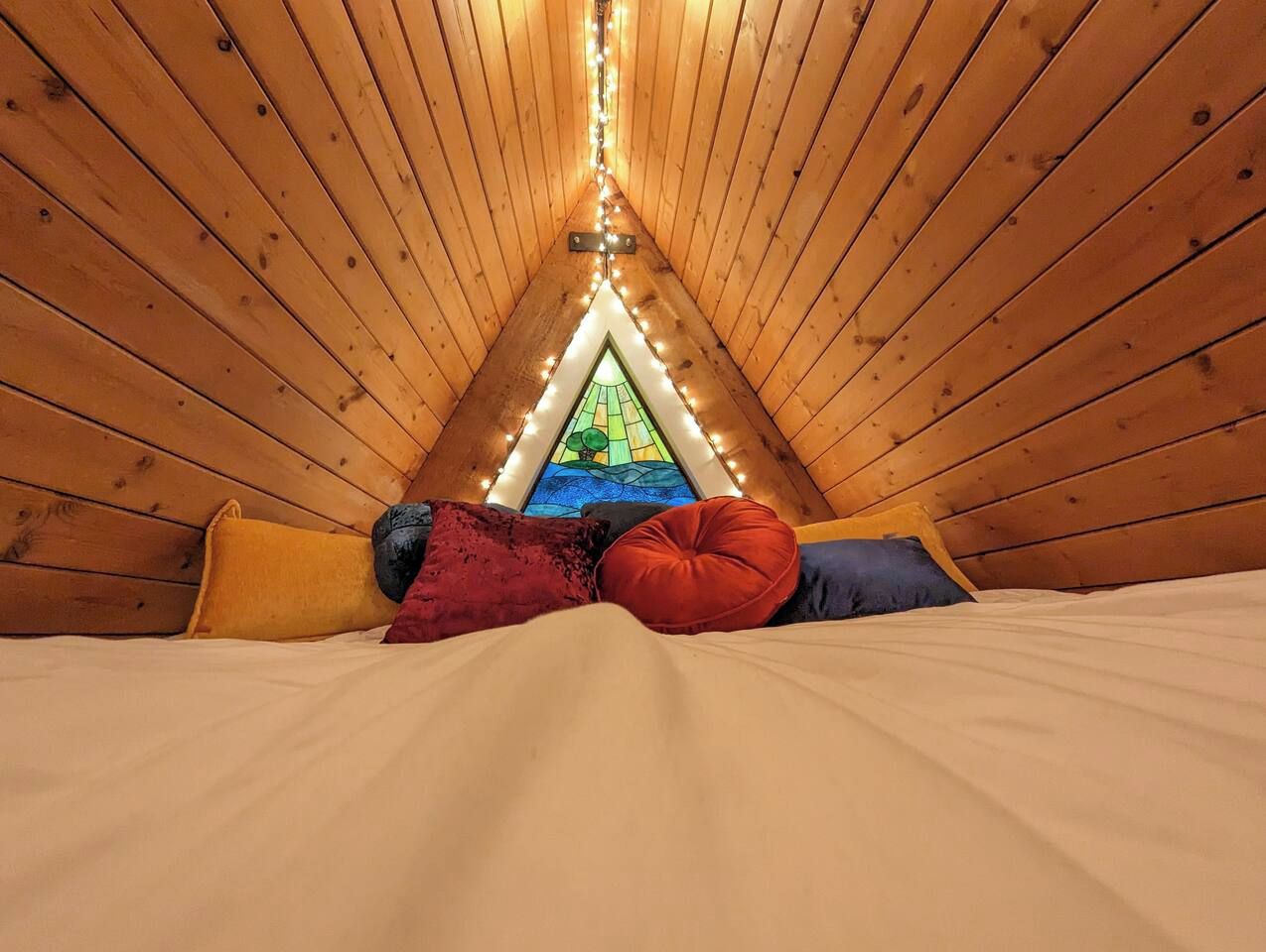 Quarto de sótão aconchegante, com teto inclinado, cama com almofadas e vista para uma paisagem verde pela janela.