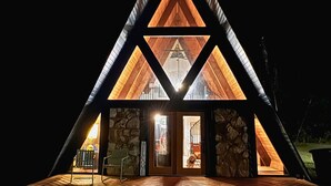 A triangular wooden cabin with a stone facade, a wooden deck, and a glass door leading to the interior. - Cozy A-Frame Cabin | Lake Access + Fire Pit (Guntersville)