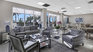 A spacious living area with a sofa, chairs, and a coffee table. There are large windows with a view of palm trees and a building. - Breezy Beachfront Condo | Panoramic Beach View (Marco Island)