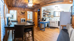 Interior acogedor de una cabaña con una mesa de comedor de madera, sillas, una zona de cocina con estanterías y un ventilador de techo.