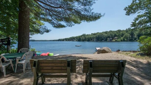Vista sobre un lago desde un banco de madera y una mesa con sillas, con vista a un lago tranquilo y una orilla boscosa.