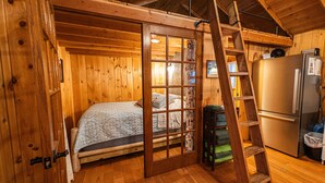 A wooden cabin interior with a bed, a ladder, and a refrigerator.