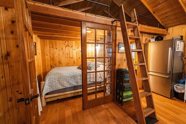 A wooden cabin interior with a bed, a ladder, and a refrigerator. - Lakeside Cabin on Beach Pond with Sauna and Beach Access (Voluntown)