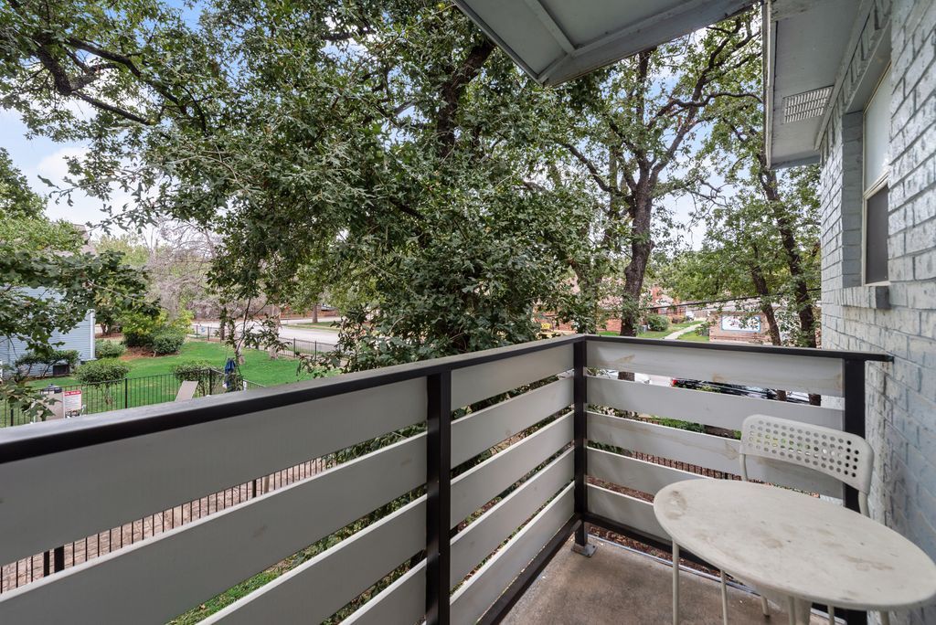 A balcony with a white table and chair, a metal railing, and a view of a green garden and trees.