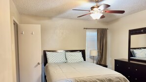 Un dormitorio con cama, ventilador de techo, ventana con persianas, mesita de noche y cómoda.