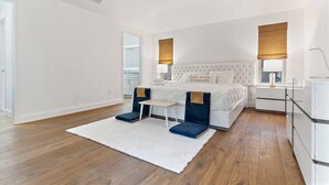 Une chambre avec un lit blanc, deux chaises bleues, un tapis blanc et un ventilateur de plafond.