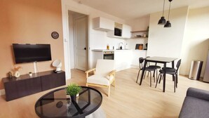 A modern living room with a TV, a round glass table, and a sofa. There is a kitchen area with a dining table and chairs. - Le Contemporain (Lens)