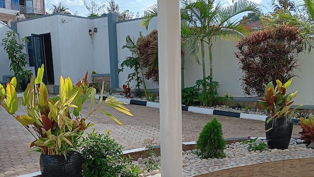 A covered outdoor area with tiled flooring, potted plants, and a white building in the background. - La Cafecito Bistro and accommodation  (Kigali)