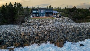 A modern house with a stone foundation, surrounded by a rocky shoreline and a forested area. - Majestic Oceanfront Chalet - Hot tub and Sauna- Private (South Shore)