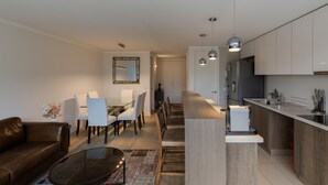 Uma sala moderna com um sofá, uma mesa de centro de vidro e uma área de jantar com cadeiras e uma mesa. Há uma cozinha com bancada e armários.