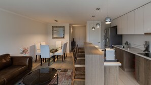 Uma sala moderna com um sofá, uma mesa de centro de vidro e uma área de jantar com cadeiras e uma mesa. Há uma cozinha com bancada e armários.