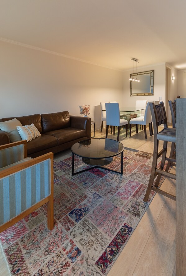 Un salón con un sofá de cuero marrón, una mesa de centro de vidrio y una alfombra estampada. Hay dos sillones y una mesa de comedor con sillas.